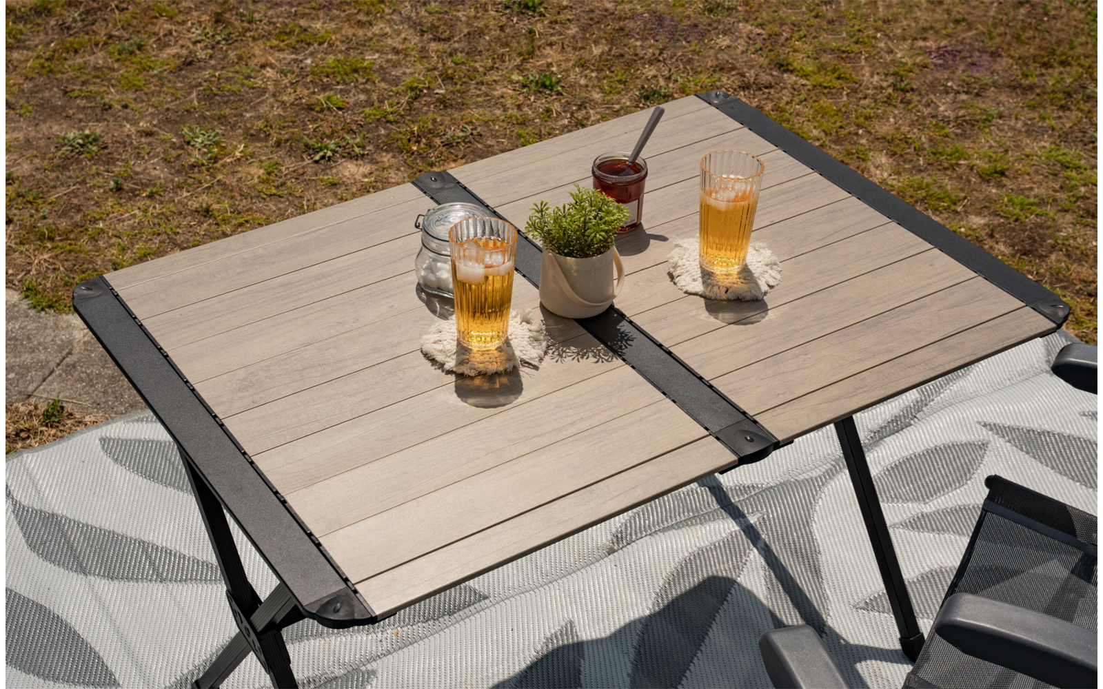 Kleiner klappbarer Tisch mit hellen Holzlatten und schwarzem Rahmen; darauf zwei Gläser mit braunem Getränk und Strohhalmen, ein weißer Blumentopf mit Pflanze und ein Glasgefäß; gemusterte Decke und Rasen im Hintergrund
