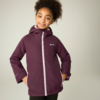 Mädchen mit hochgebundenem lockigem Haar trägt eine bordeauxfarbene Kapuzenjacke mit weißem Reißverschluss und Logo auf der Brust, Hände in den Taschen, heller Hintergrund