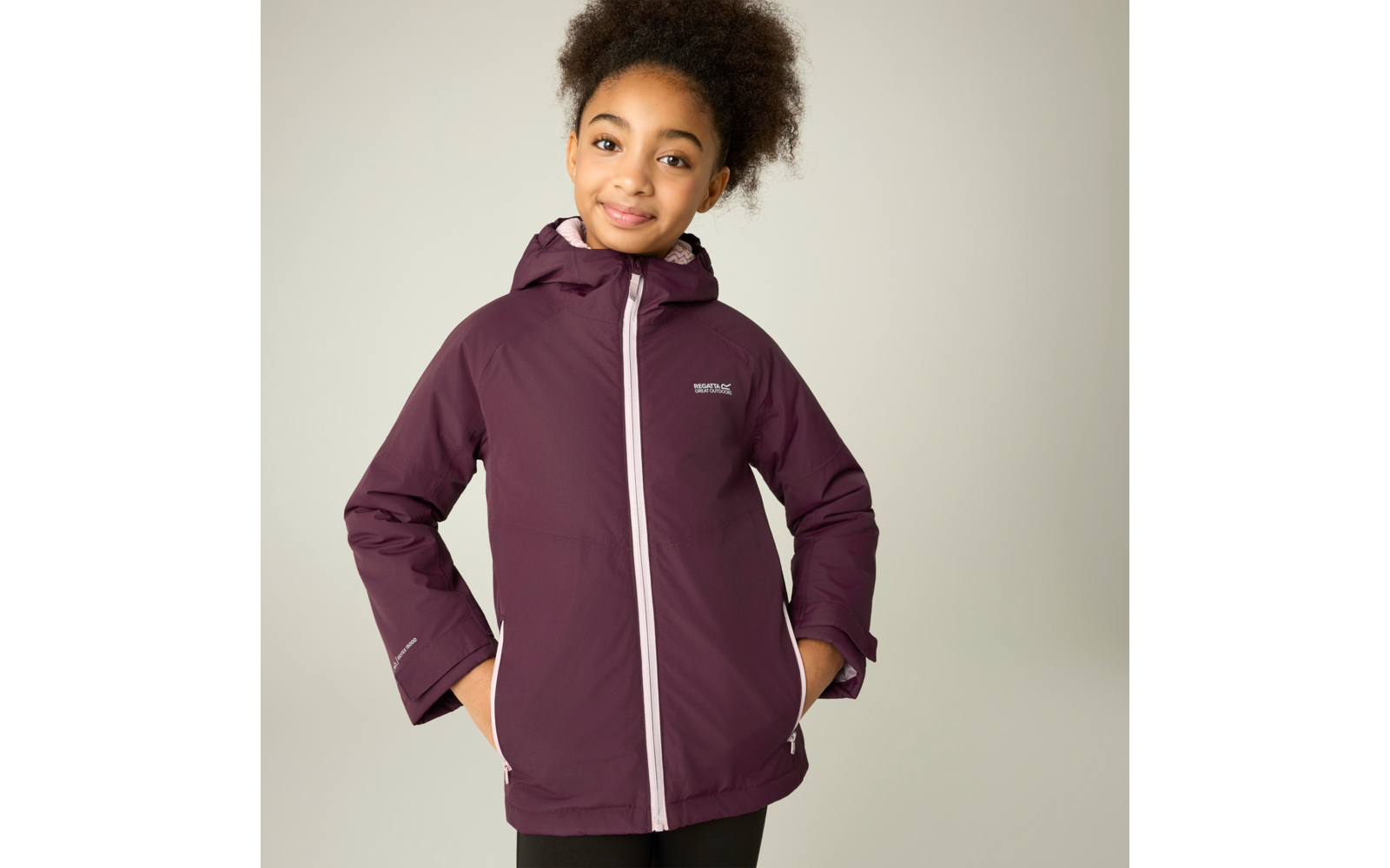 Mädchen mit hochgebundenem lockigem Haar trägt eine bordeauxfarbene Kapuzenjacke mit weißem Reißverschluss und Logo auf der Brust, Hände in den Taschen, heller Hintergrund