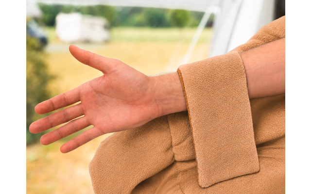 Uitgestrekte onderarm en hand naast een mouw van een beige jas; wazig grasveld en een caravan op de achtergrond