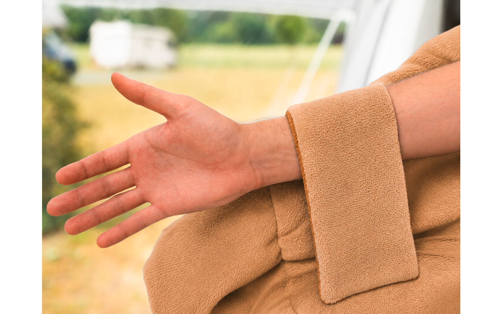 Uitgestrekte onderarm en hand naast een mouw van een beige jas; wazig grasveld en een caravan op de achtergrond