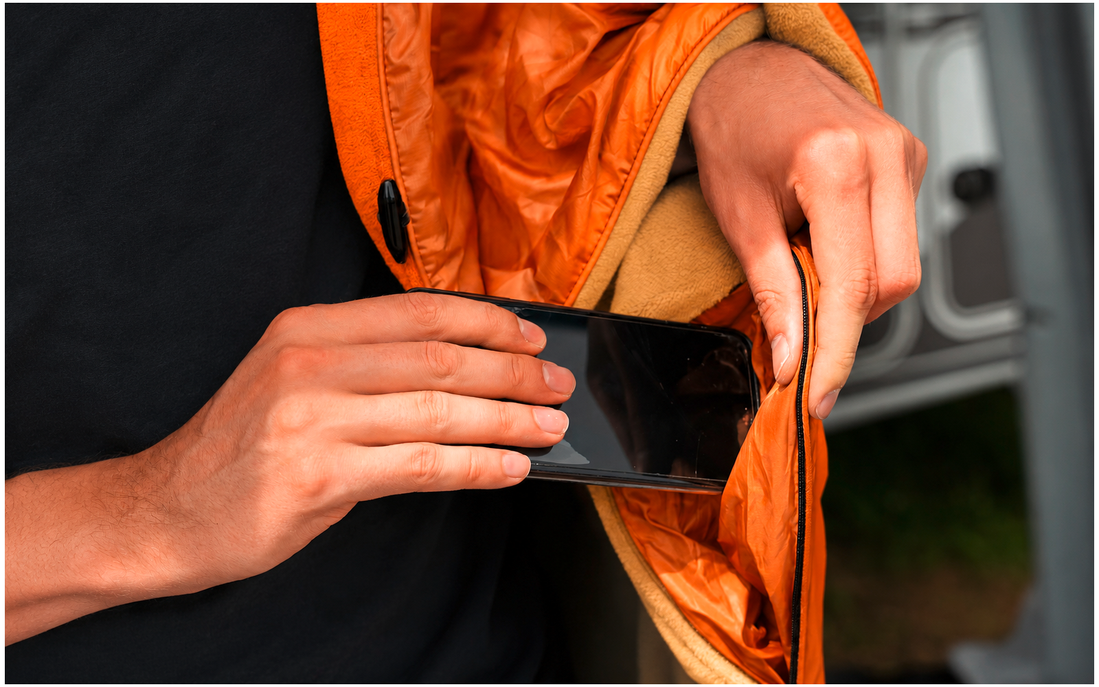 Twee handen steken een smartphone in de binnenzak van een oranje jas, zwart shirt zichtbaar