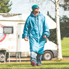Persoon in lange blauwe gewatteerde jas, muts, zonnebril, spijkerbroek en rode schoenen, houdt een beker vast, loopt op gras voor een witte camper, berkenstam op de achtergrond