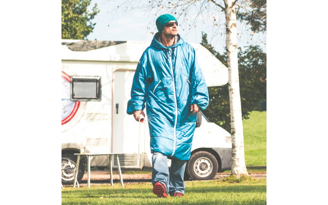 Persoon in lange blauwe gewatteerde jas, muts, zonnebril, spijkerbroek en rode schoenen, houdt een beker vast, loopt op gras voor een witte camper, berkenstam op de achtergrond