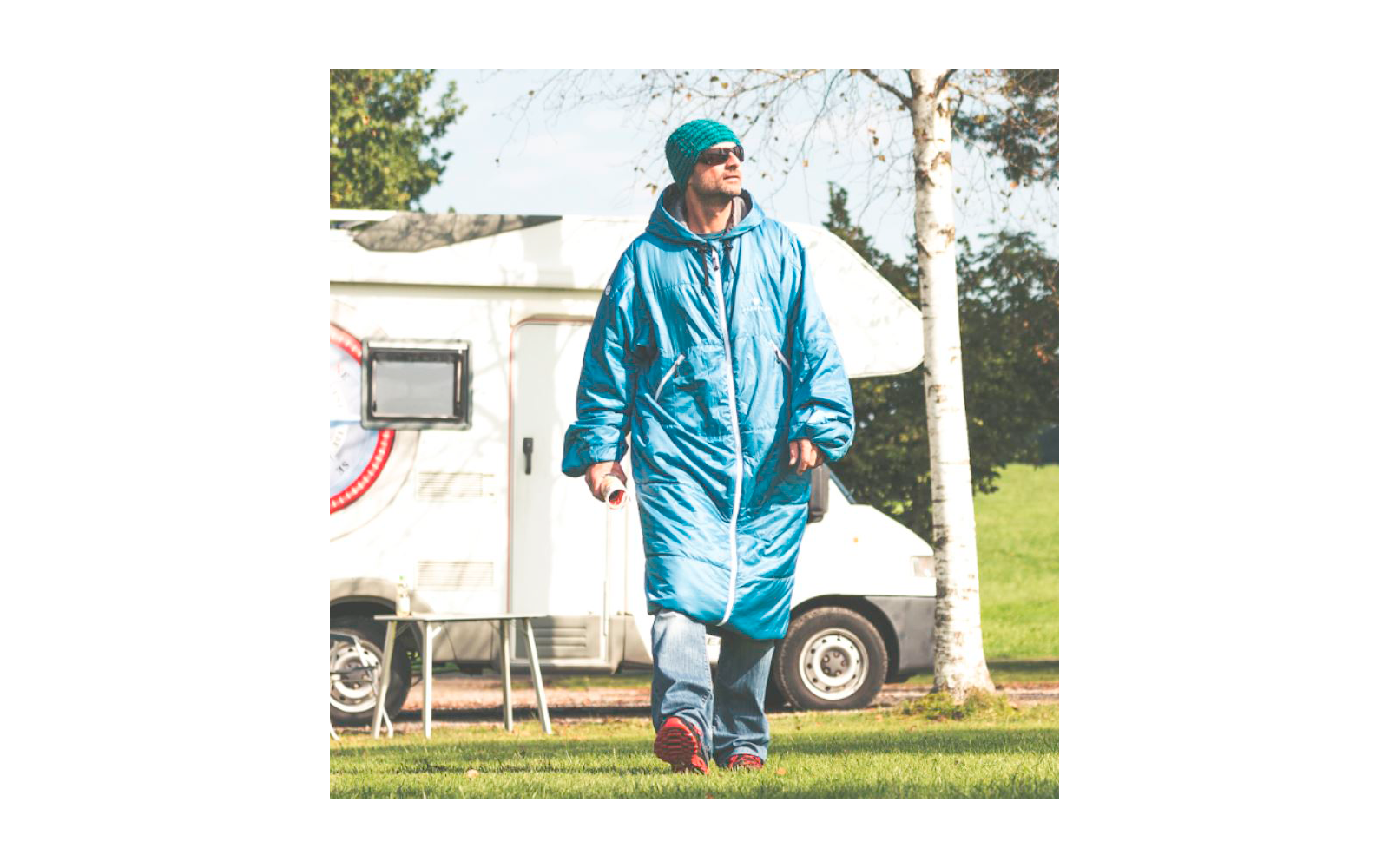 Persoon in lange blauwe gewatteerde jas, muts, zonnebril, spijkerbroek en rode schoenen, houdt een beker vast, loopt op gras voor een witte camper, berkenstam op de achtergrond