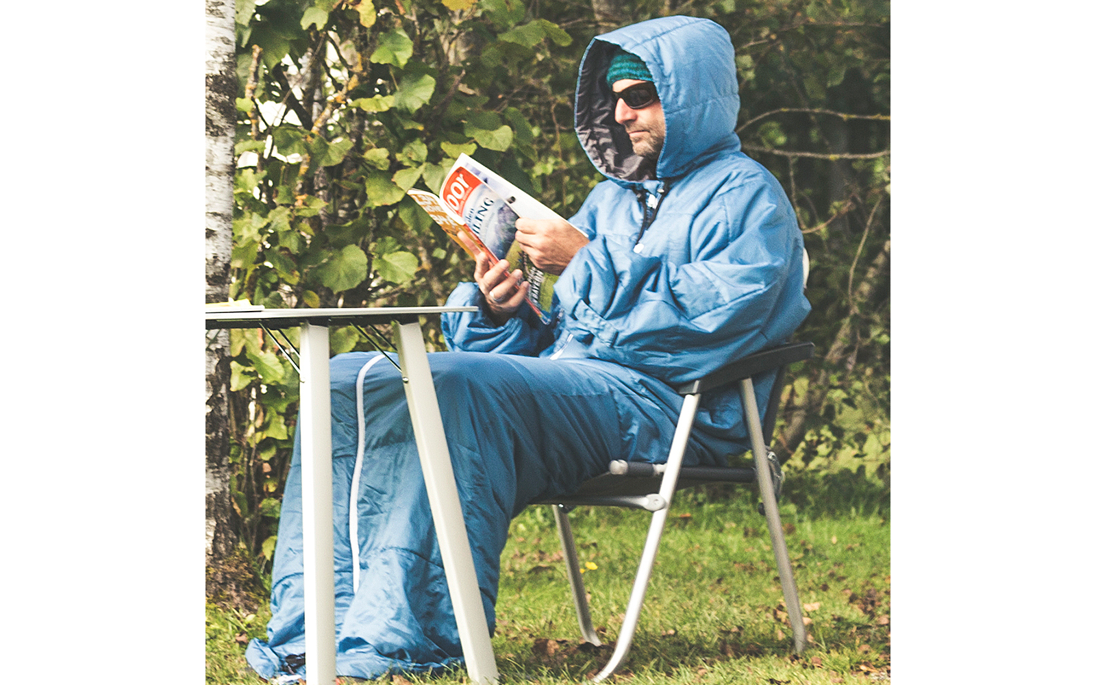 Persoon zit op een vouwstoel buiten, draagt een blauwe capuchonjas en zonnebril, leest een tijdschrift; witte tafel ervoor, gras en bomen op de achtergrond
