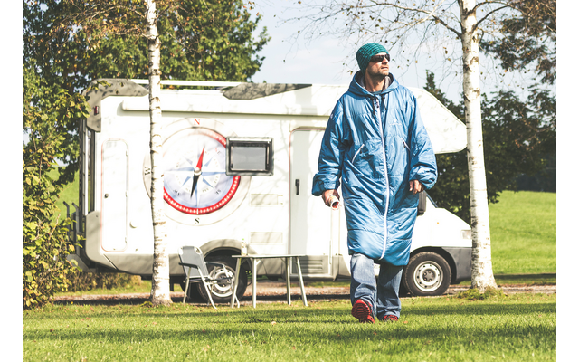 Persoon in blauwe slaapzak-achtige jas, groene muts en zonnebril loopt op gras voor een camper met kompaslogo, naast een campingtafel en stoelen