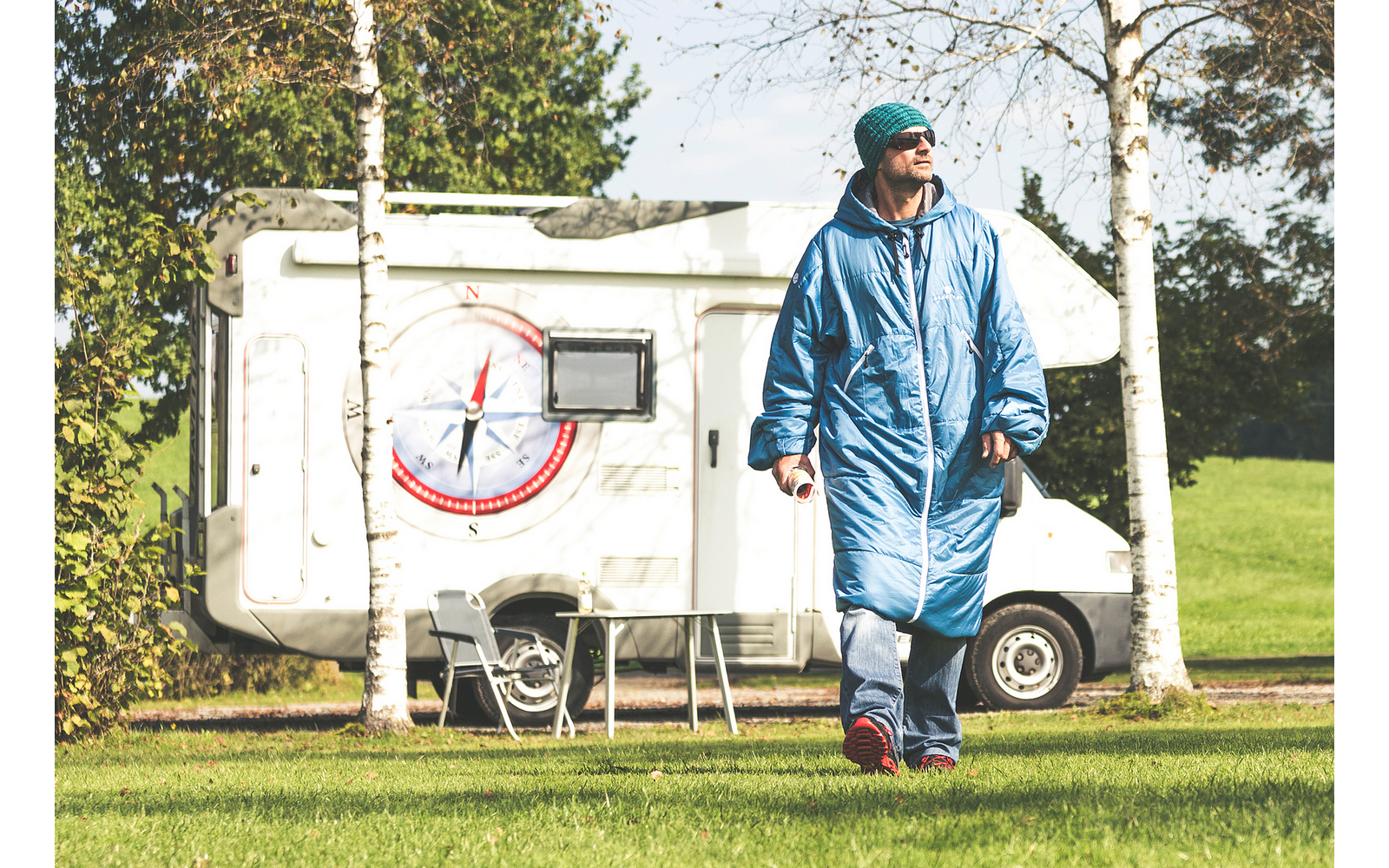 Persoon in blauwe slaapzak-achtige jas, groene muts en zonnebril loopt op gras voor een camper met kompaslogo, naast een campingtafel en stoelen