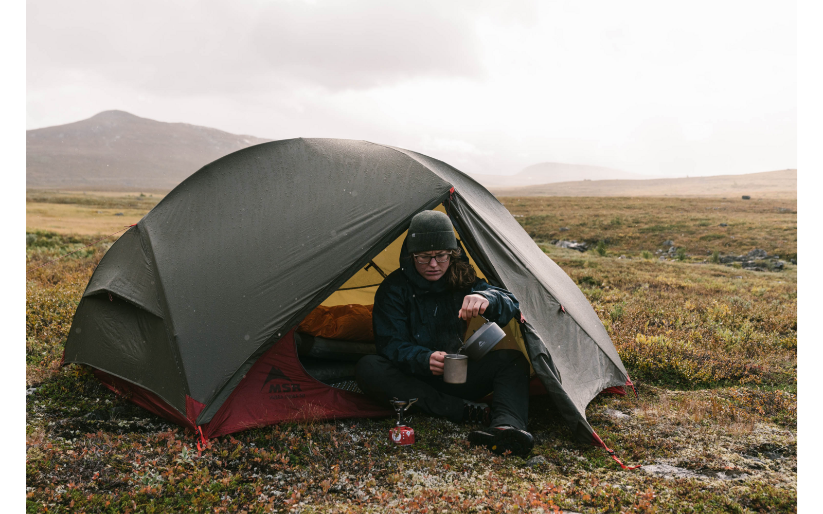 Person sitzt vor einem grünen Zelt in einer weiten, grasbewachsenen Landschaft mit Hügeln im Hintergrund
