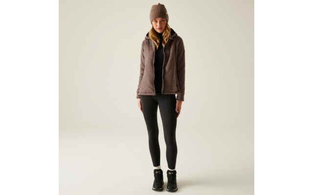 Eine Frau trägt eine braune Mütze, einen grauen Pullover, eine braune Weste, schwarze Leggings und schwarze Schuhe