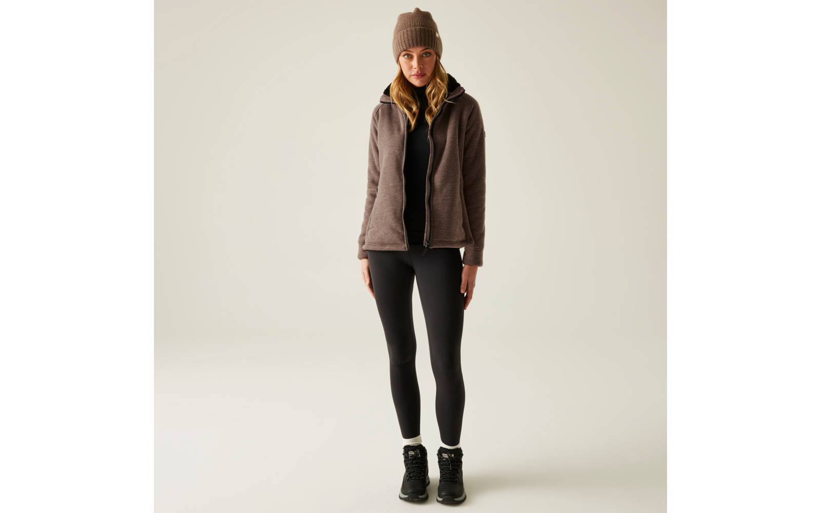 Eine Frau trägt eine braune Mütze, einen grauen Pullover, eine braune Weste, schwarze Leggings und schwarze Schuhe