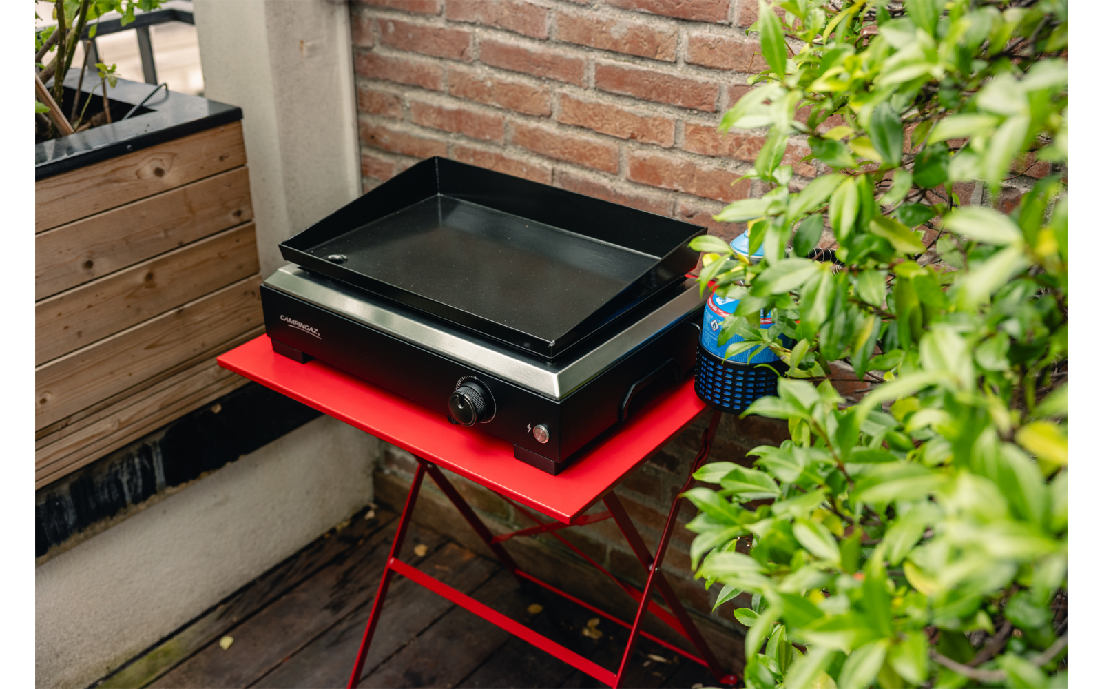 Rechteckiges schwarzes Gerät mit flacher Metalloberfläche und Drehknopf auf einem roten Klapptisch auf einem Balkon; Ziegelwand im Hintergrund; links Holzkasten mit Pflanzen; rechts grüne Blätter