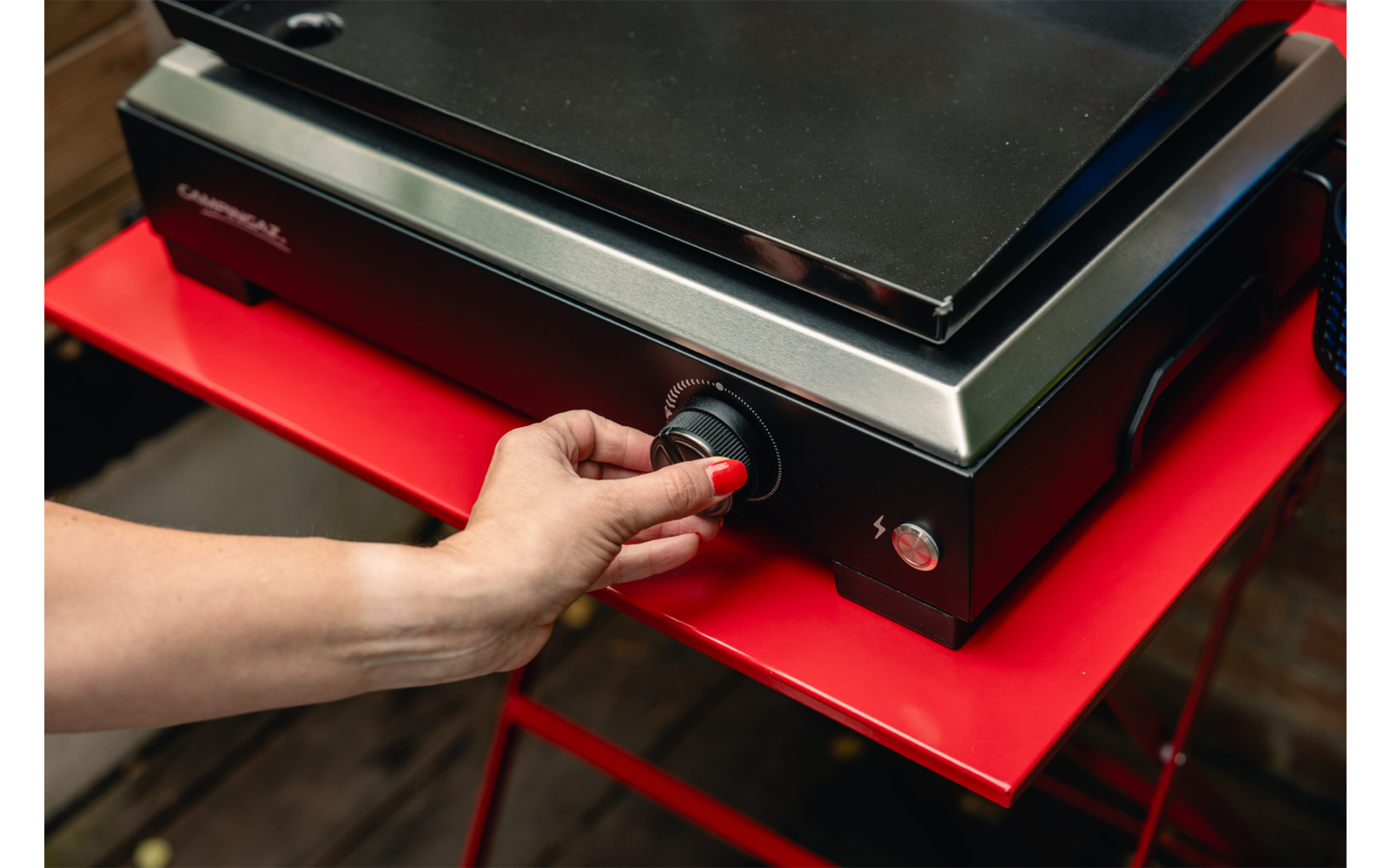 Hand dreht schwarzen Drehknopf an schwarzem Elektrogrill mit Edelstahlfront; Gerät steht auf rotem Tisch