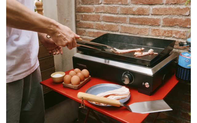 Hände mit Zange wenden Speckstreifen auf einer Grillplatte; daneben Eierschachtel, kleine Schale, Teller mit rohem Speck, Spachtel auf rotem Tisch vor Ziegelwand
