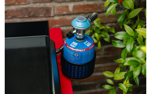 Blaue Gaskartusche mit Regler und schwarzem Schlauch auf roter Fläche, Ziegelwand im Hintergrund, grüne Blätter rechts