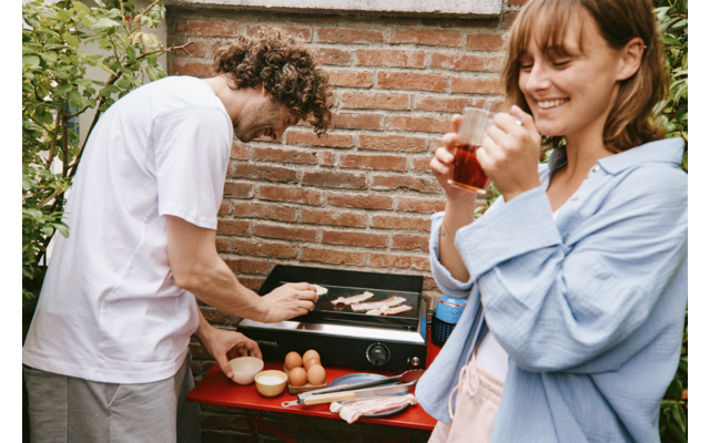 Zwei Personen neben einem Tischgrill; eine Person wendet Speckstreifen mit einer Zange, die andere hält ein Glas und lächelt; Ziegelwand im Hintergrund