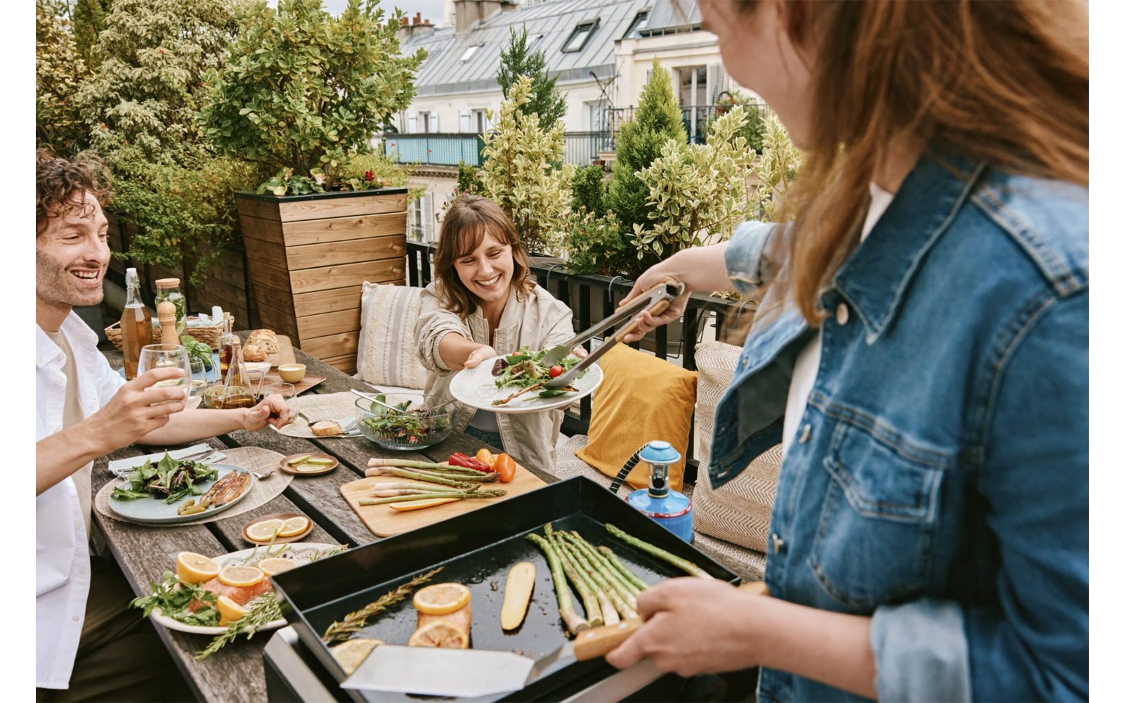 Drei Personen an einem Außentisch; eine Person reicht mit Zangen Salat; auf dem Tisch Teller, Getränke und gegrilltes Gemüse auf einer Grillplatte; Pflanzen und Gebäude im Hintergrund