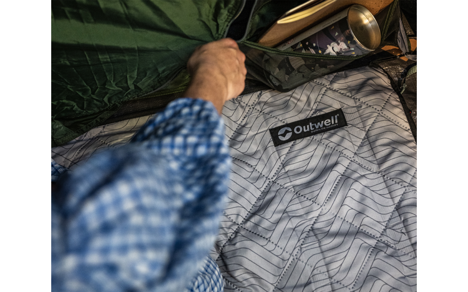 Hand in blau kariertem Hemdsärmel öffnet Reißverschluss eines grünen Schlafsacks, hellgraues Innenfutter mit Linienmuster und Aufnäher 'Outwell', goldfarbene Tasse daneben