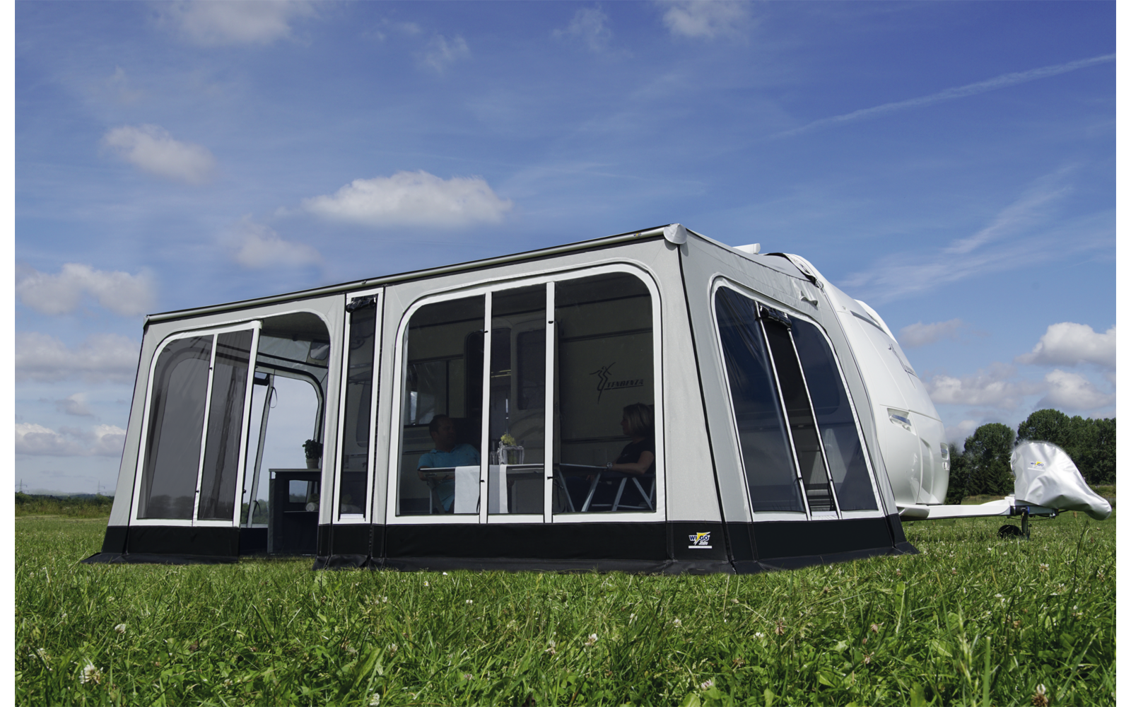 Wohnwagen mit angebautem, transparentem Vorzelt auf einer grünen Wiese unter blauem Himmel