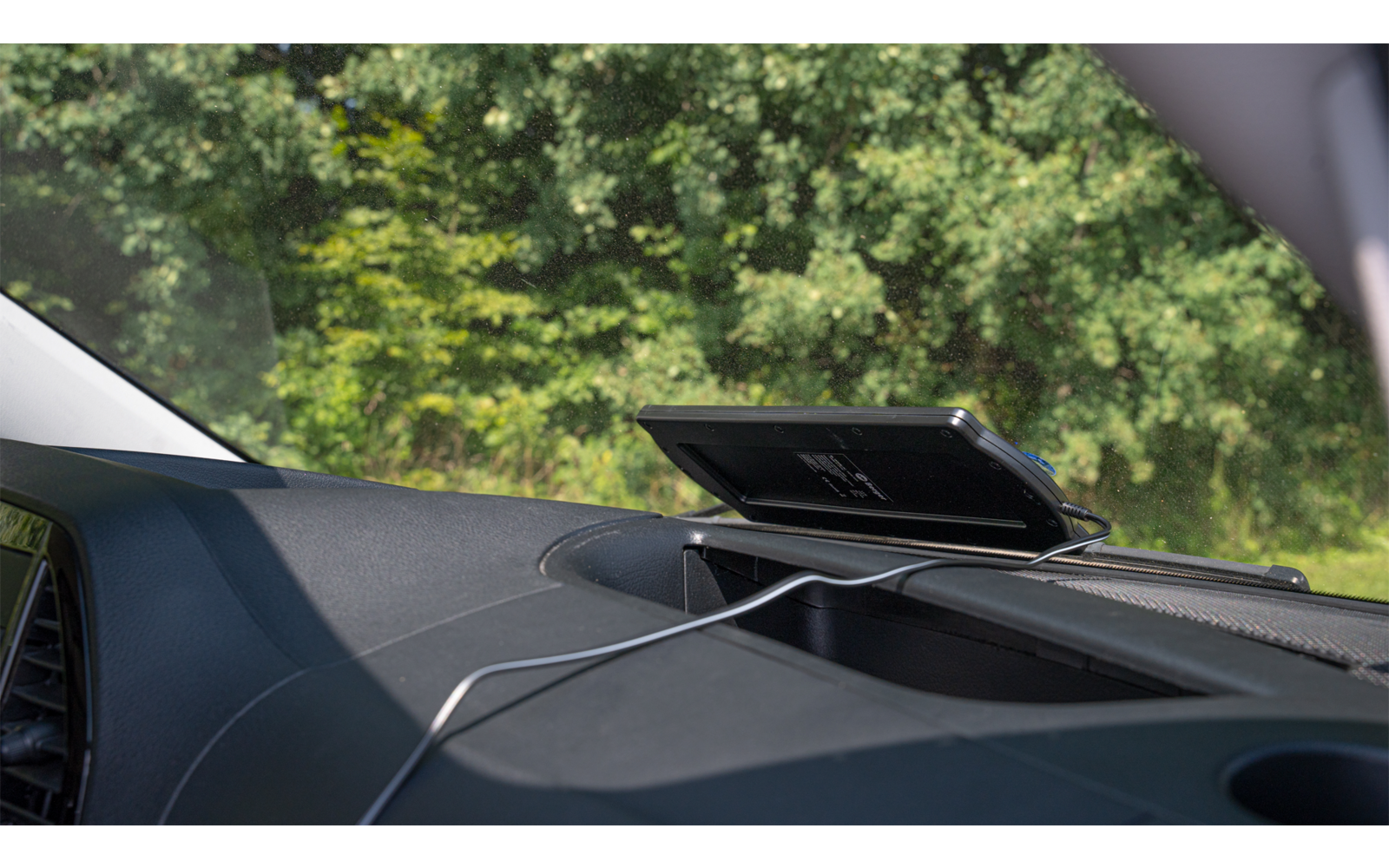 Blick durch die Windschutzscheibe eines Autos auf einen digitalen Tachometer und eine grüne Baumkulisse im Hintergrund