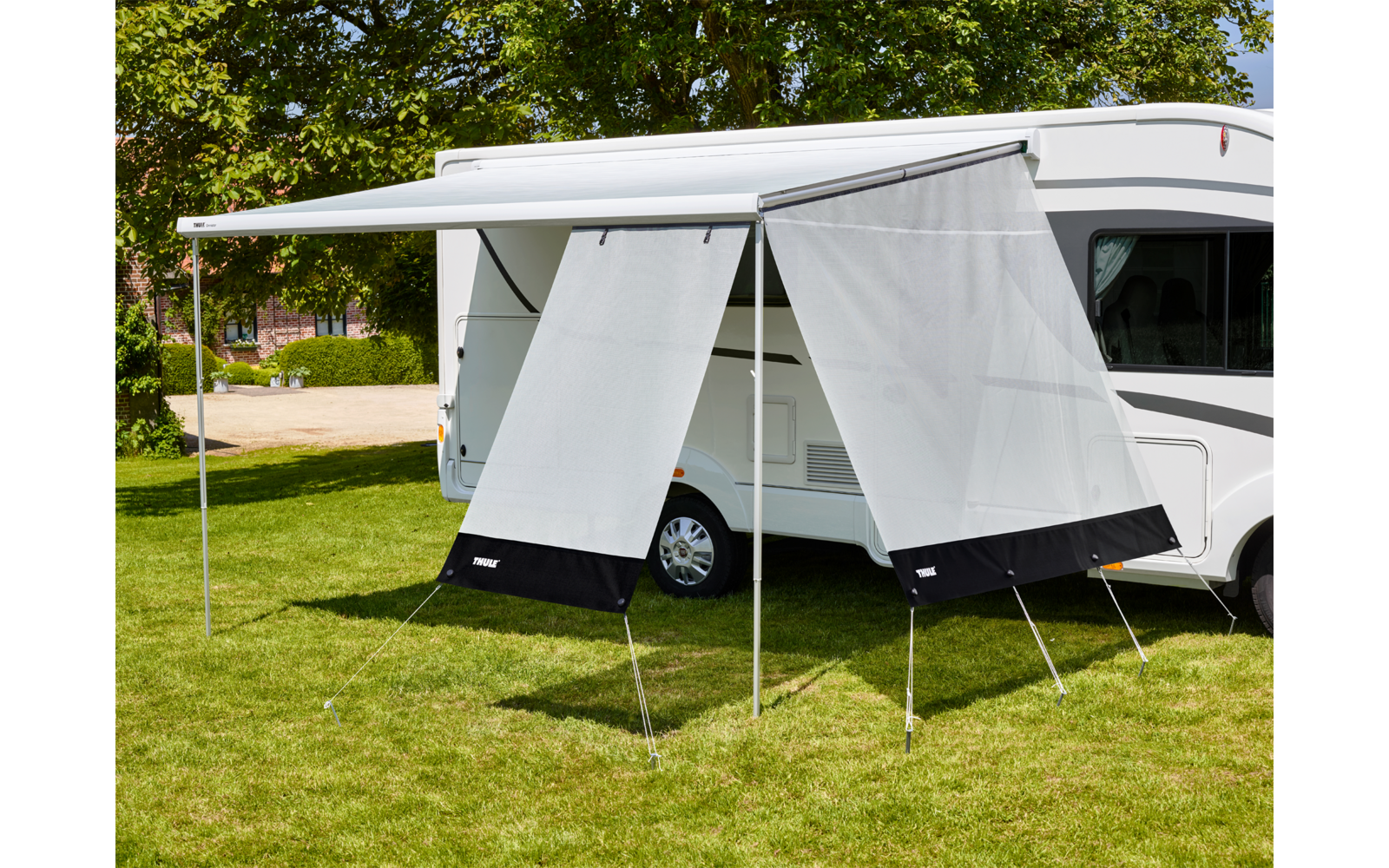 Camper bianco su prato con tenda estesa e due pannelli laterali in tessuto su pali metallici, alberi sullo sfondo