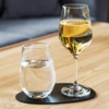 Weinglas mit gelblicher Flüssigkeit und Trinkglas mit klarem Wasser auf schwarzer ovaler Unterlage auf Holztisch, unscharfe Couch im Hintergrund
