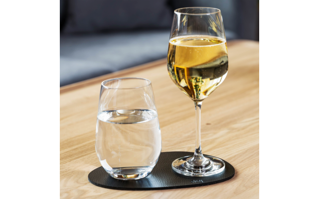 Weinglas mit gelblicher Flüssigkeit und Trinkglas mit klarem Wasser auf schwarzer ovaler Unterlage auf Holztisch, unscharfe Couch im Hintergrund