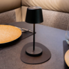 Schwarze Tischlampe auf rundem schwarzem Untersetzer auf einem Tisch, zwei gelbe Keramikteller, Besteck, Trinkglas und gepolsterte Sitzbank im Hintergrund
