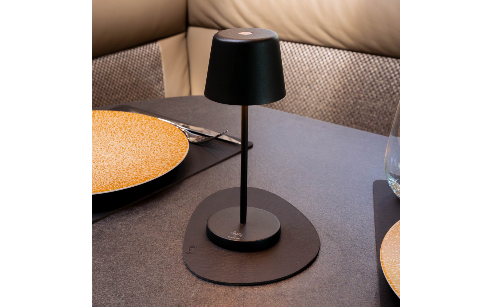 Schwarze Tischlampe auf rundem schwarzem Untersetzer auf einem Tisch, zwei gelbe Keramikteller, Besteck, Trinkglas und gepolsterte Sitzbank im Hintergrund