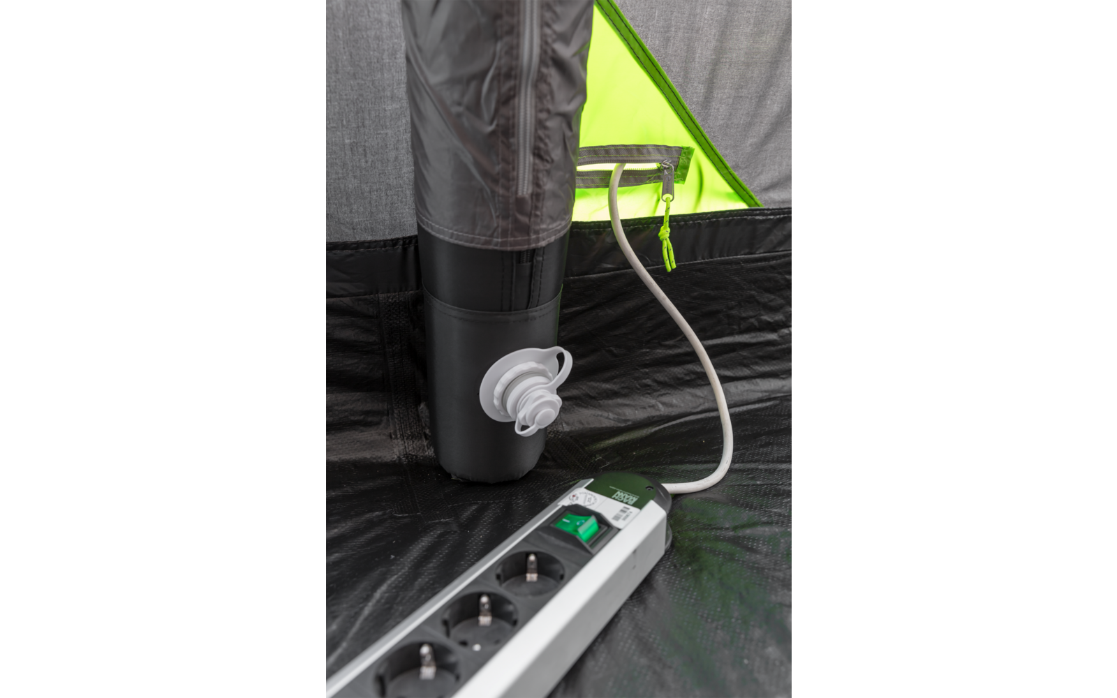 Interieur van een tent met een zwarte opgeblazen buis met een wit ventiel, een witte kabel en een stekkerdoos met een groene schakelaar op een zwarte vloer; op de achtergrond een felgroen stoffen paneel met ritssluiting