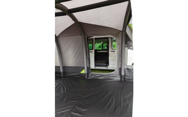 Interieur van een grijze luifel met zwarte vloer; door een opening is een witte bus met open schuifdeur en groene bomen op de achtergrond zichtbaar