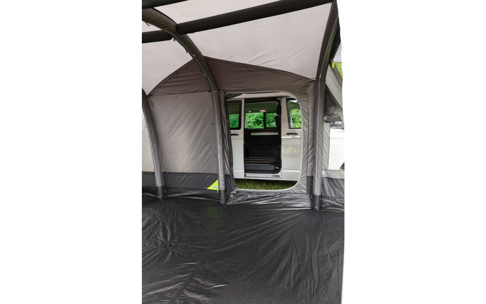 Interieur van een grijze luifel met zwarte vloer; door een opening is een witte bus met open schuifdeur en groene bomen op de achtergrond zichtbaar