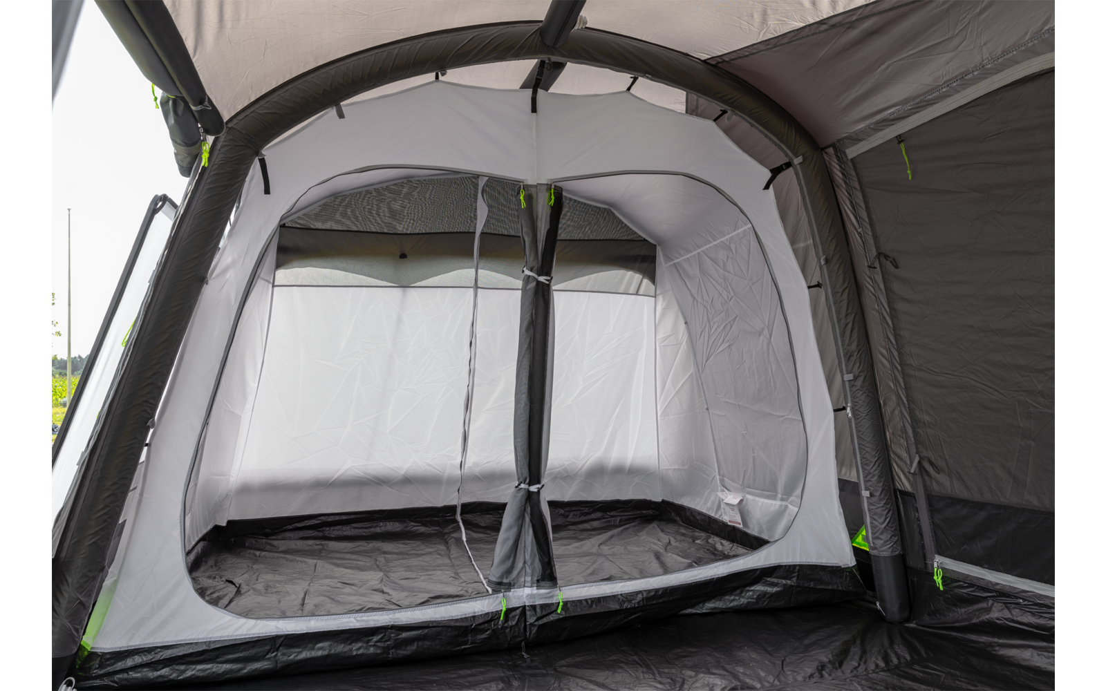 Binnenkant van een grijze tent met lichtgrijze binnenwanden, zwart grondzeil, grote rechthoekige opening met ritsen, gebogen buizen/stangen en groene ritsaccenten