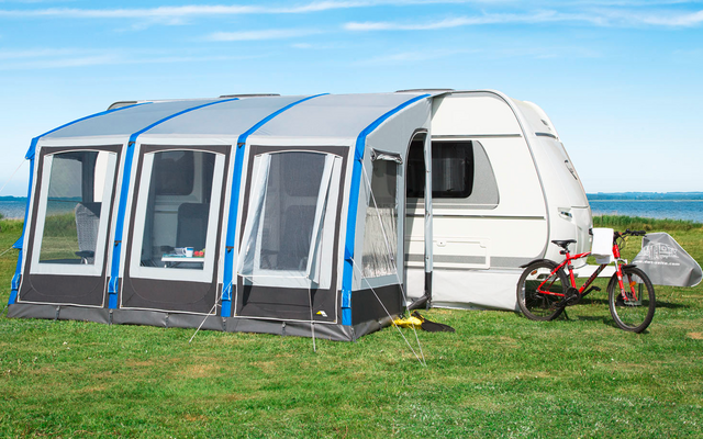 Caravane avec auvent bleu-gris attaché sur une prairie, deux vélos au premier plan, mer et ciel bleu en arrière-plan
