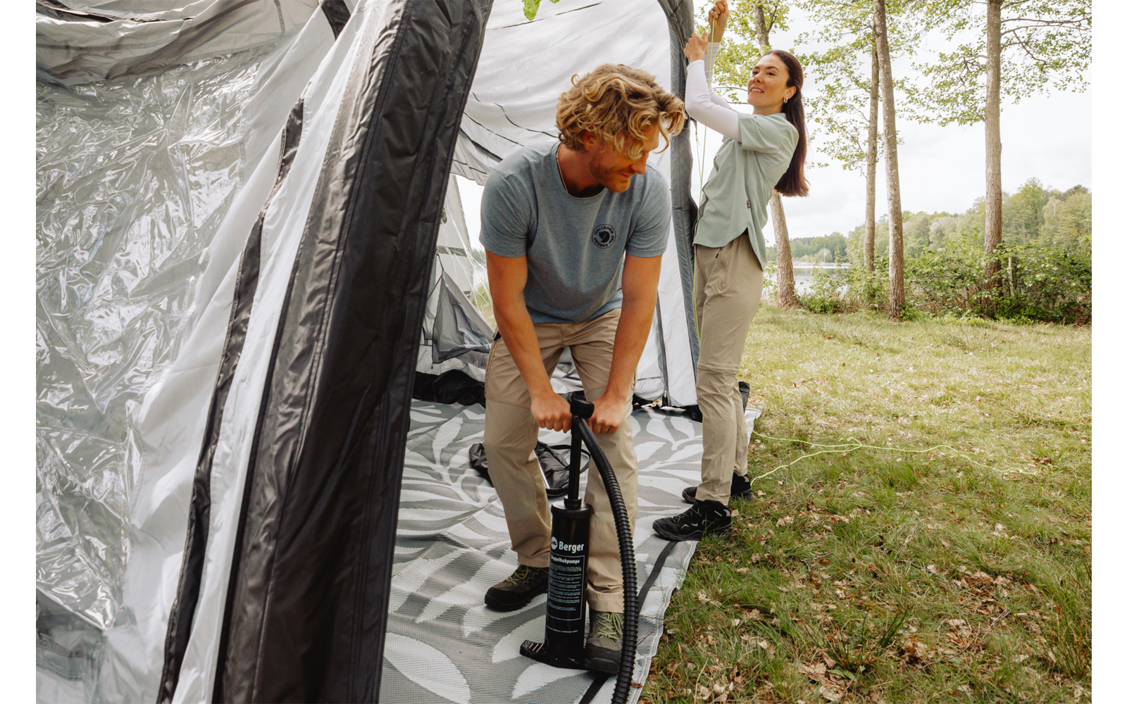 Two people assembling an inflatable tent; one operates a hand pump at the tent, the other adjusts the outer fabric; grass and trees in the background