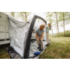 Two people attach an inflatable awning to a caravan on a grassy area with trees
