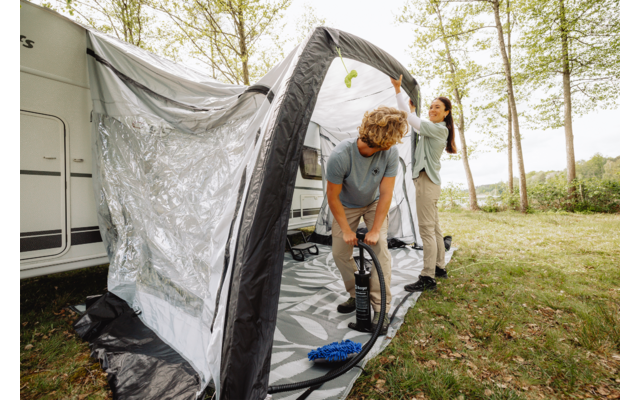 Two people attach an inflatable awning to a caravan on a grassy area with trees