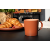 Terrakottafarbene Tasse auf dunklem Tisch; im Hintergrund Teller mit Croissants und Beeren, geflochtener Korb und unscharfer Herd