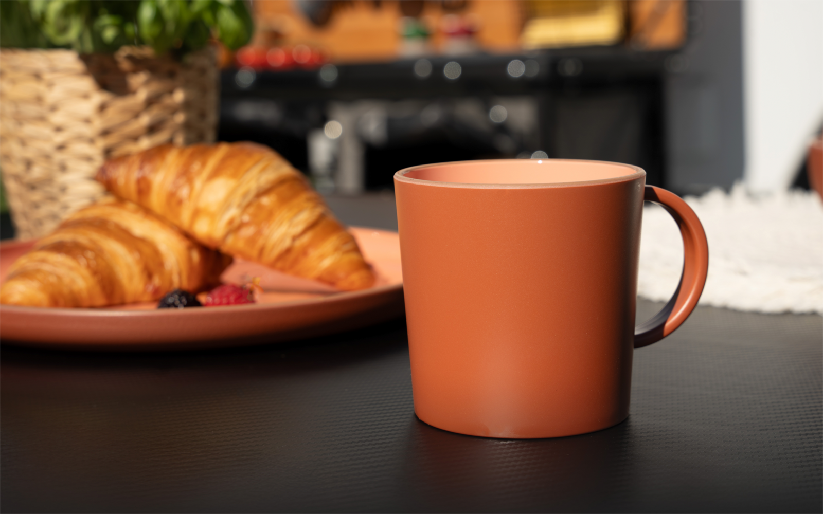 Terrakottafarbene Tasse auf dunklem Tisch; im Hintergrund Teller mit Croissants und Beeren, geflochtener Korb und unscharfer Herd