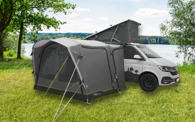 Graues Wohnmobil mit angehängtem grauen Zelt auf Wiese neben See und Bäumen