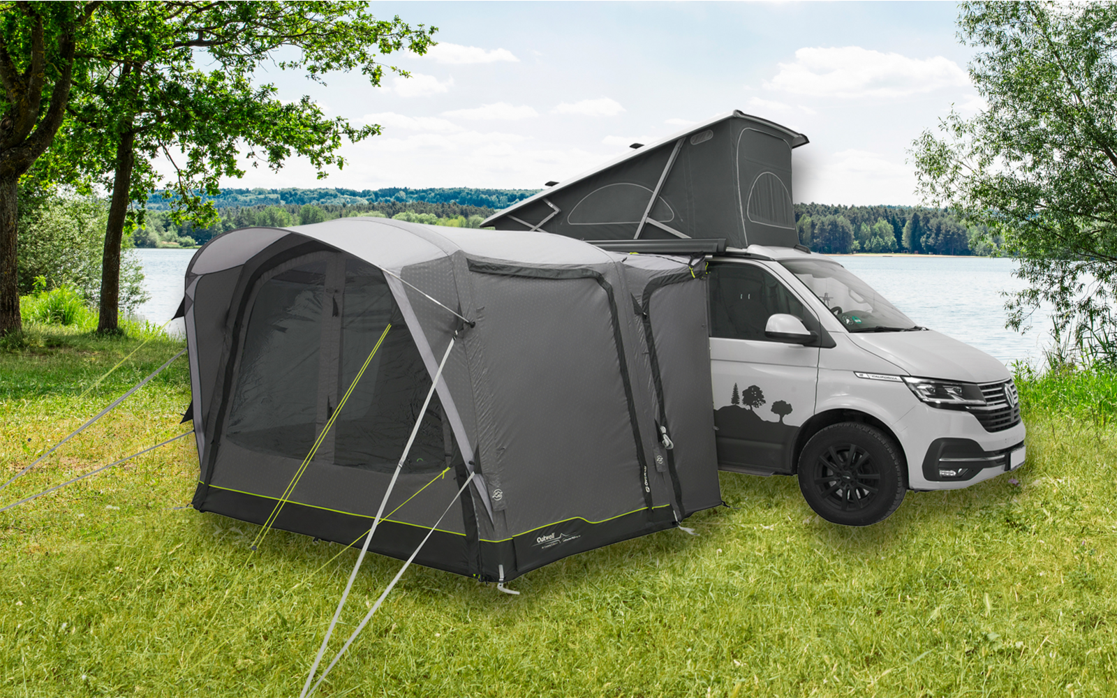 Graues Wohnmobil mit angehängtem grauen Zelt auf Wiese neben See und Bäumen