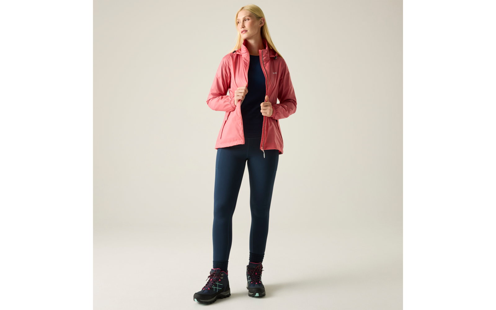 Person mit blondem Haar steht vor hellem Hintergrund, trägt pinke Jacke, dunkle Leggings und schwarze Wanderschuhe, hält die Jacke am Reißverschluss und blickt zur Seite