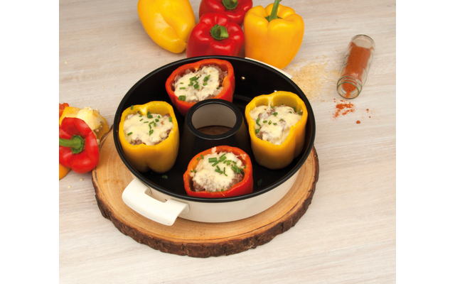 Vier rote und gelbe Paprikahälften mit weißer Füllung und gehackten grünen Kräutern in einer runden Auflaufform auf einer Holzscheibe; im Hintergrund drei ganze Paprika und ein umgekipptes Gewürzglas