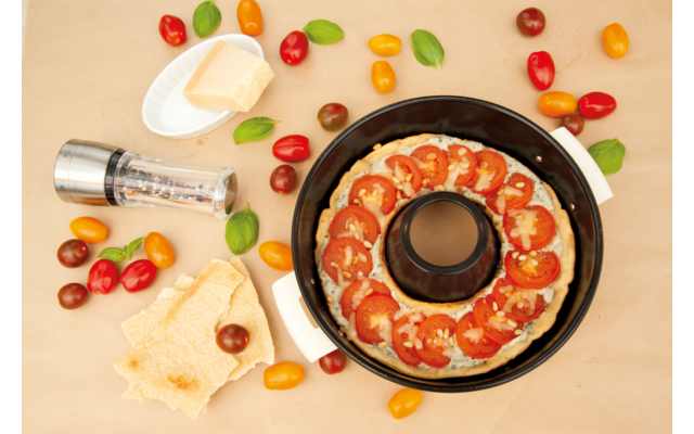 Runde Backform mit Teig und Tomatenscheiben, umgeben von gelben und roten Kirschtomaten, Basilikumblättern, einer Pfeffermühle, einem Käsekeil auf einem Teller und Brotstücken auf beigem Hintergrund
