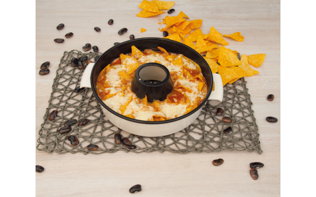 Runde weiße Auflaufform mit schwarzem Rand und schwarzem Mittelring, gefüllt mit geschmolzenem Käse und roter Sauce, gelbe Tortilla-Chips darauf und daneben, dunkle Bohnen auf grauem geflochtenem Tischset auf hellem Holztisch