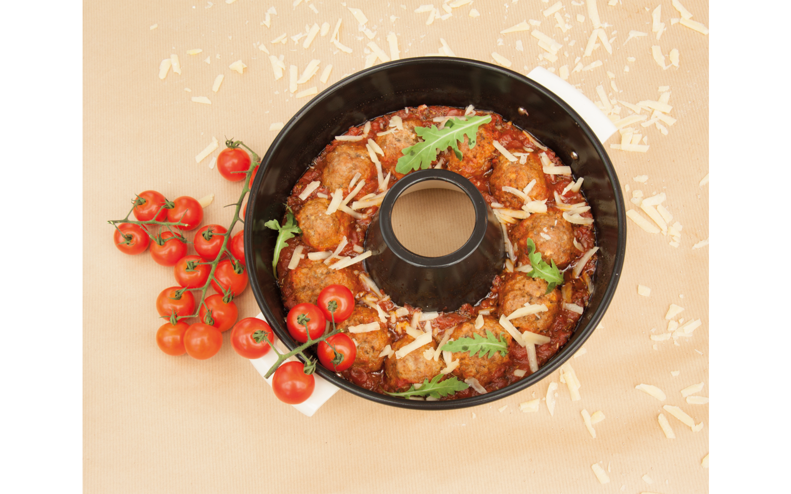 schwarze Kranzbackform mit runden braun-roten Bällchen in roter Sauce, bestreut mit geriebenem Käse und einigen grünen Blättern; Trauben-Kirschtomaten links auf hellem Untergrund