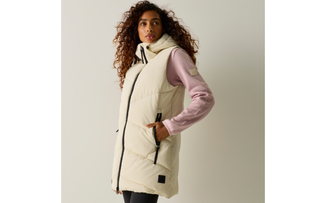 Persona con gilet imbottito lungo crema con cappuccio sopra un maglione rosa, mani nelle tasche, capelli ricci, guarda verso destra