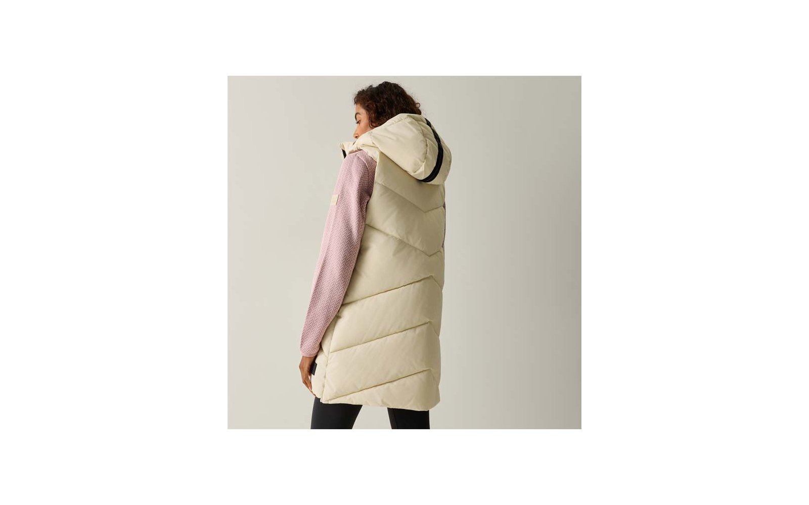 Persona vista di spalle che indossa un lungo gilet trapuntato crema con cappuccio, maglione rosa e leggings neri, sfondo chiaro uniforme