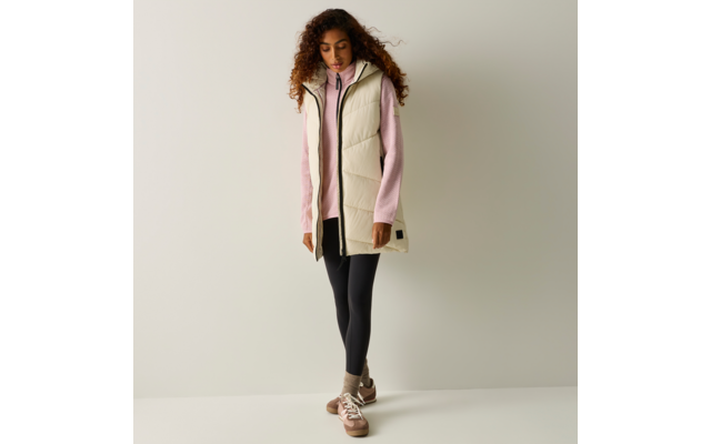 Persona con capelli ricci in piedi davanti a una parete chiara, indossa un gilet imbottito color crema, un maglione rosa, leggings neri e sneakers marroni, guarda verso il basso