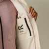 Mano con unghie curate tiene aperta una giacca beige chiaro, fodera interna con tasca interna con zip, logo nero 'R' e piccolo testo sulla tasca, piping rosa, maglione rosa a motivo sotto, sfondo neutro